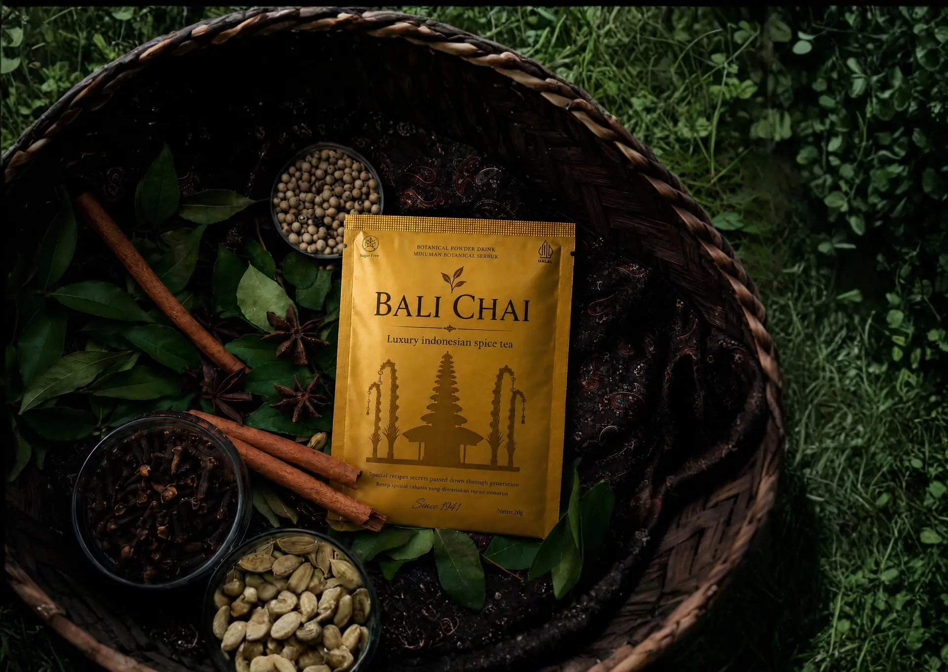 Top-down view of a Bali Chai pouch in a woven basket surrounded by cloves, cinnamon, cardamom and fresh leaves